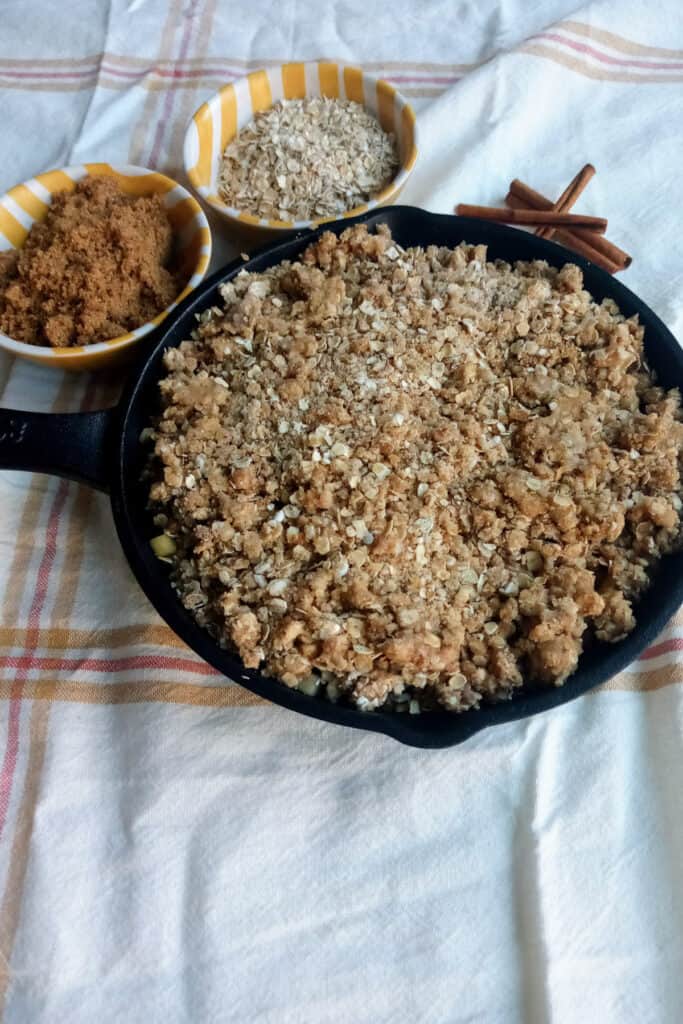 Baked apple crisp next to three cinnamon sticks and a bowl of oats and brown sugar.