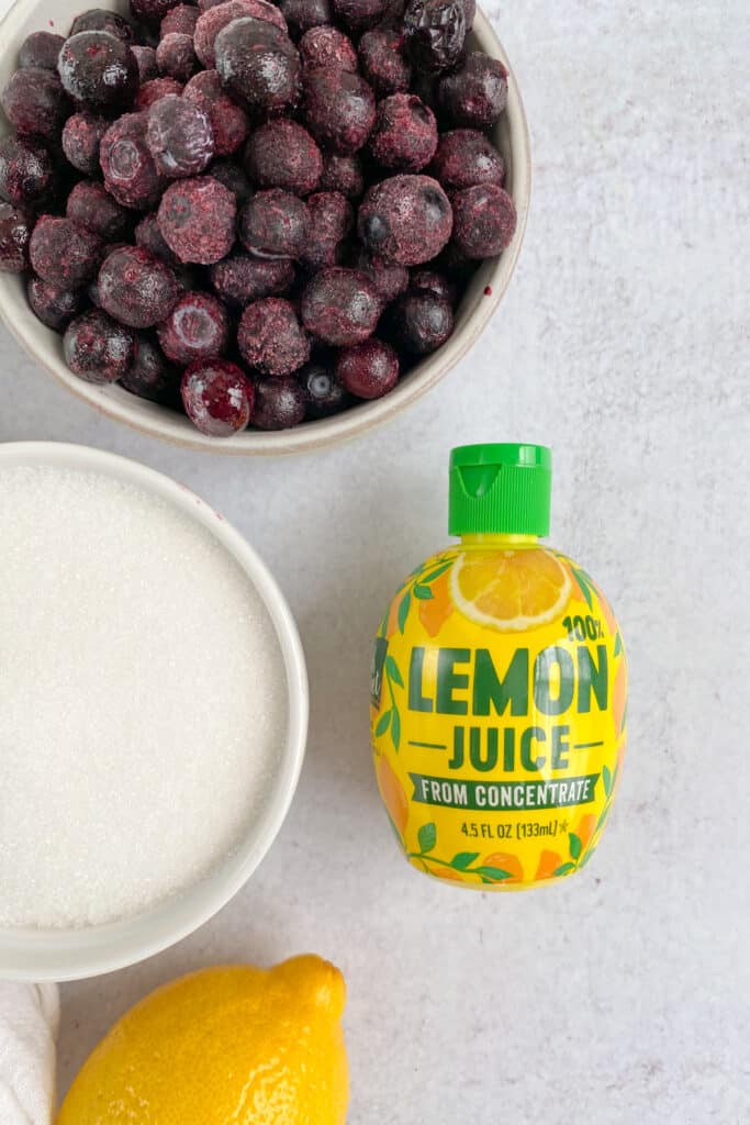 Bowls of frozen blueberries and sugar and a lemon-shaped bottle of lemon juice.