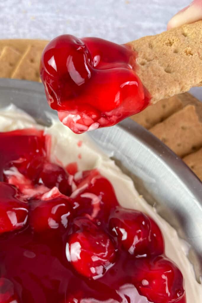 A person pulling a graham cracker out of the cheesecake dip with lots of dip and a large cherry on top.
