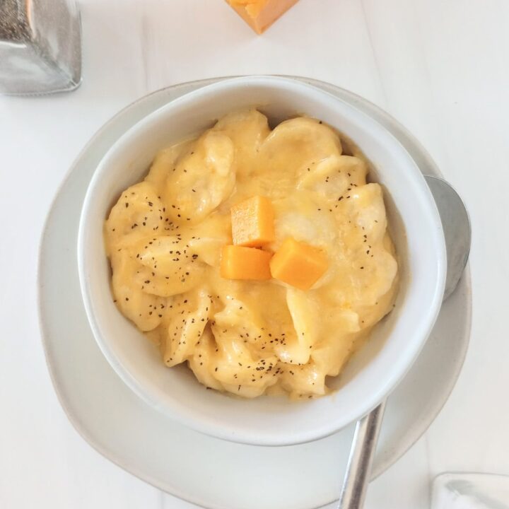 Slow cooker mac and cheese in a white bowl next to salt and pepper shakers. Topped with ground black pepper and cubed cheese.