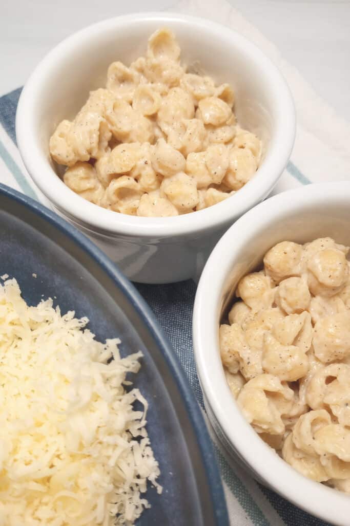 Two white bowls of mac and cheese and shredded white cheddar cheese.