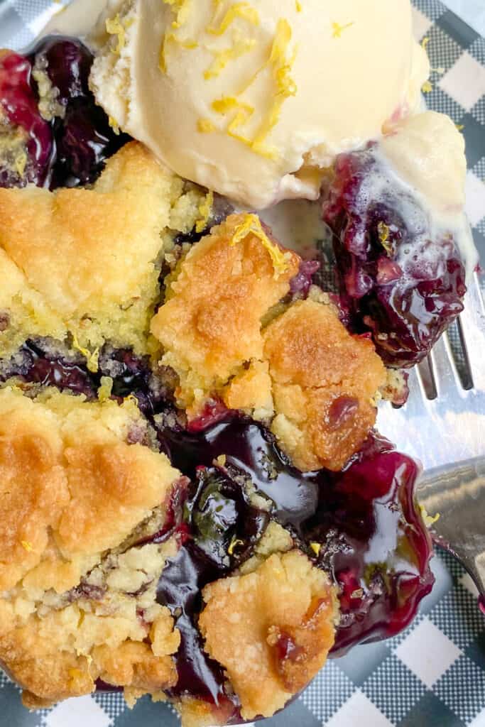 Close up picture of blueberry cobbler and ice cream on a dish where you can see the bursting blueberries and buttery soft cake mix topping.