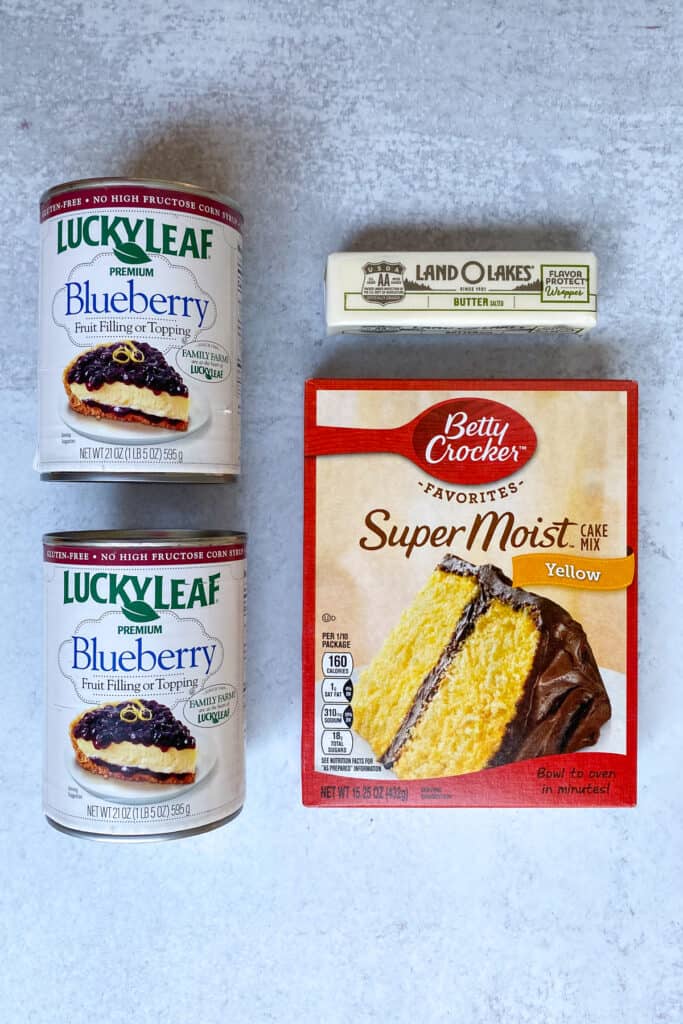 Two cans of blueberry pie filling, a boxed yellow cake mix and stick of butter.