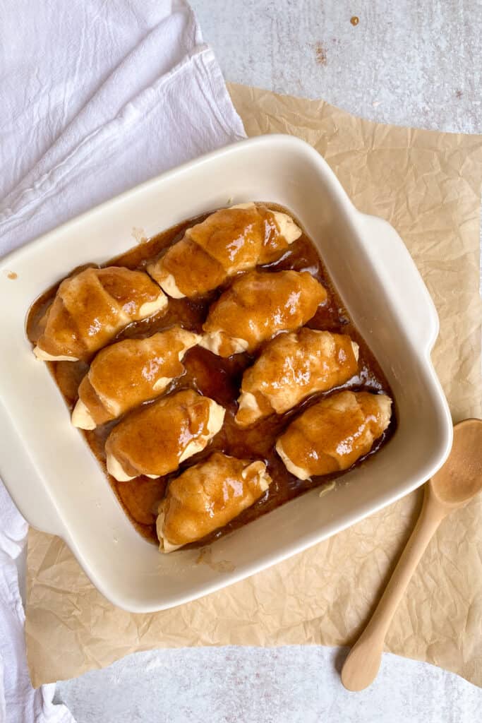 Eight peach dumplings in a baking dish with brown sugar butter topping spread over them.