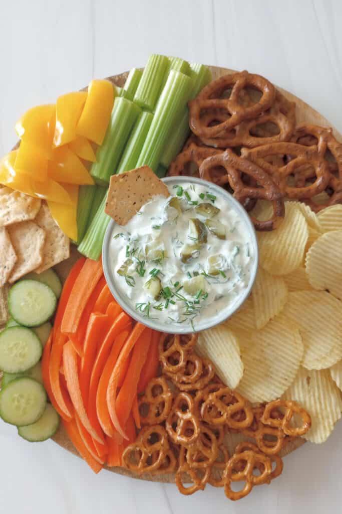 A cracker in a bowl of dill pickle dip on a circular tray loaded with fresh sliced veggies, chips, pretzels and crackers for dipping.