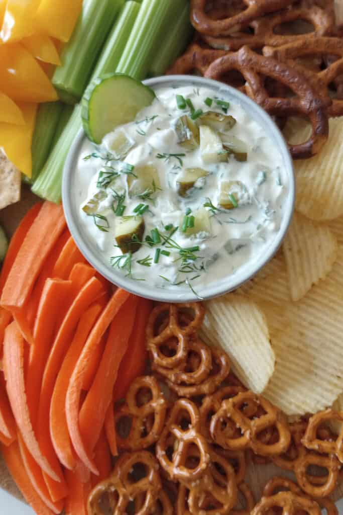 Sliced cucumber in a bowl of dill pickle dip on a tray with chopped veggies, chips and pretzels.