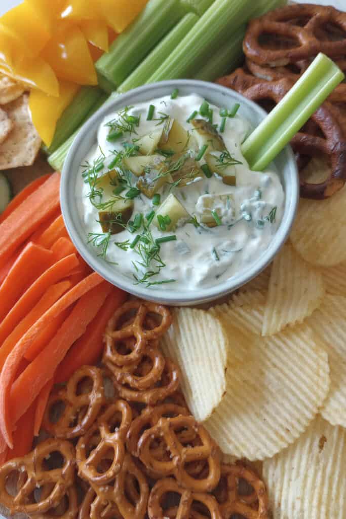 Celery stick in a bowl of dill pickle dip on a tray with veggies, pretzels and chips for dipping.
