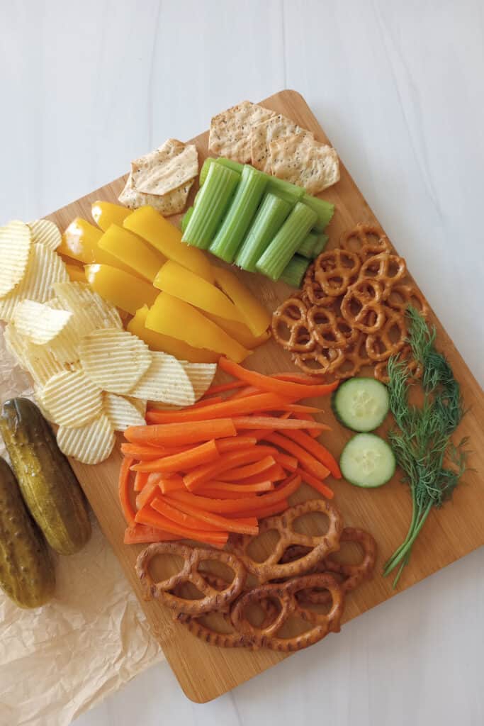 A cutting board with sliced carrots, and celery, and potato chips and crackers to use as dippers in dill pickle dip.