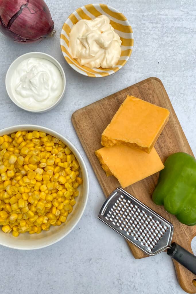 Ingredients to make Frito Corn Dip including a bowl of corn, block of cheddar cheese, red onion, green pepper, and small bowls containing sour cream and mayo.