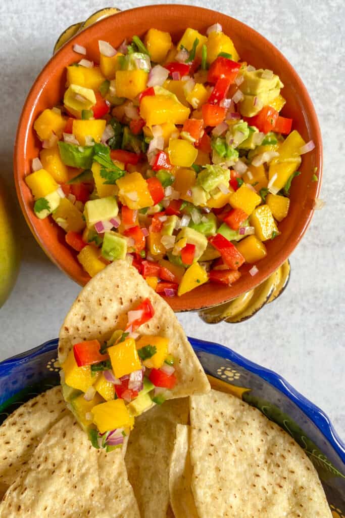 A bowl of Mango Avocado Salsa with a tortilla chip resting on the side, loaded with a scoop full of salsa.