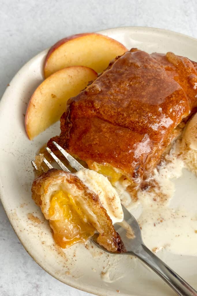 A fork with a bite of a crescent roll peach dumpling and ice cream on it.