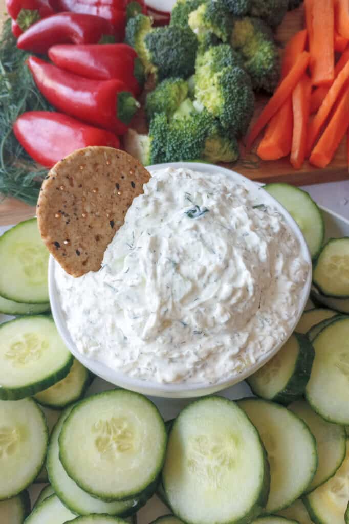 Multigrain cracker dipped in cool cucumber dip. Dip is arranged around a colorful veggie board. 