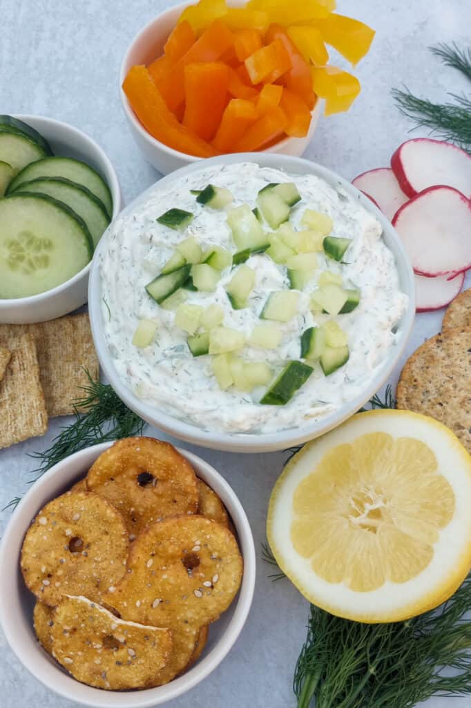 Dip topped with diced cucumber on a table served with sliced radishes, bagel chips, Triscuit crackers, and multigrain crackers. 