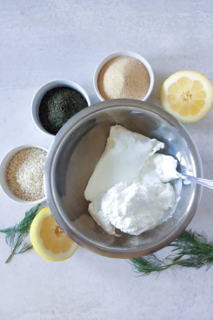 Sour cream and cream cheese in a small mixing bowl. Garlic powder, dill weed, and minced onion seasonings are arranged in small white bowls around the dip. Halved lemons are arranged around cucumber dip with fresh dill for show and a color pop.