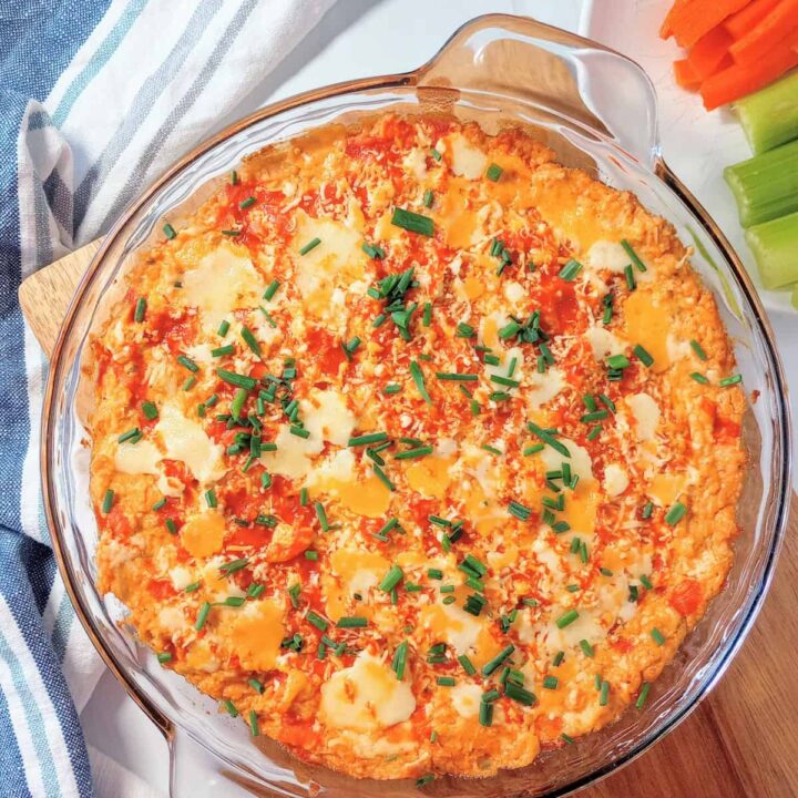 Super bowl buffalo chicken dip fresh out of the oven topped with chives. Carrot and celery sticks are arranged on a white plate next to dip.