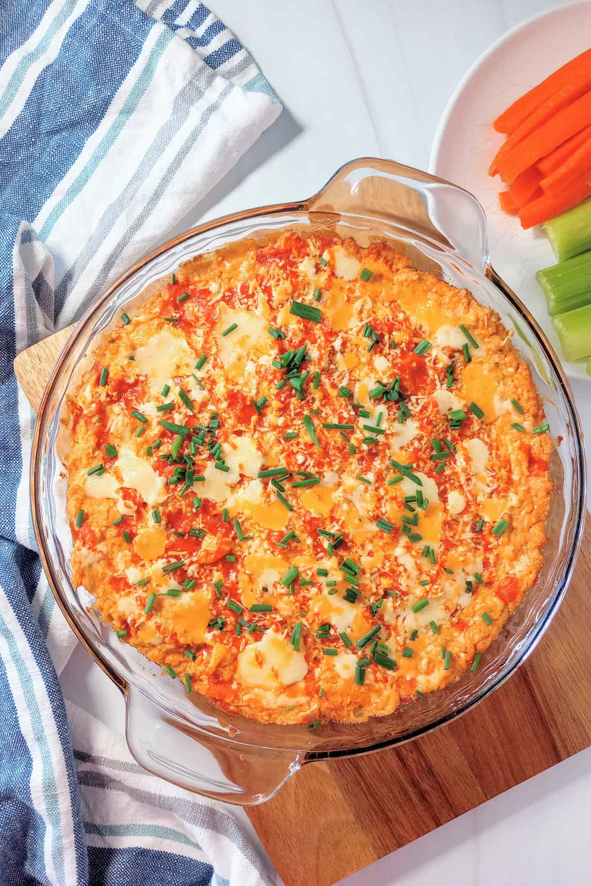 Super bowl buffalo chicken dip fresh out of the oven topped with chives. Carrot and celery sticks are arranged on a white plate next to dip.