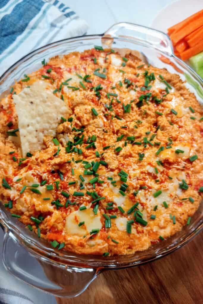 Tortilla chip dipped in warmed chicken dip set out on a table. Carrot and celery sticks are arranged next to the dip for a healthy chip option.