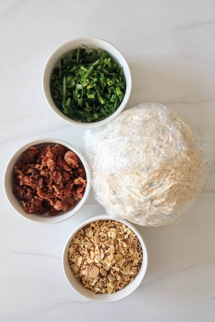 Jalapeno popper cheese ball wrapped in saran wrap next to three white bowls of crumbled bacon, roasted almonds, and chives.