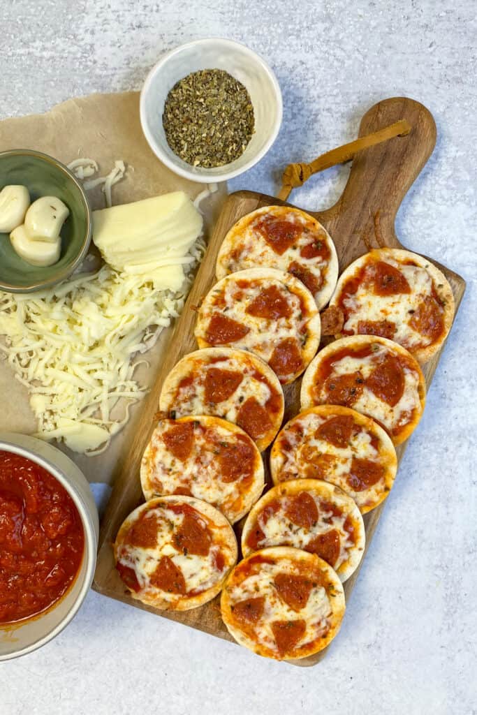 A dozen 3 inch tortilla pizzas on a serving board next to grated mozzarella cheese, a bowl of sauce and a bowl of Italian seasoning.