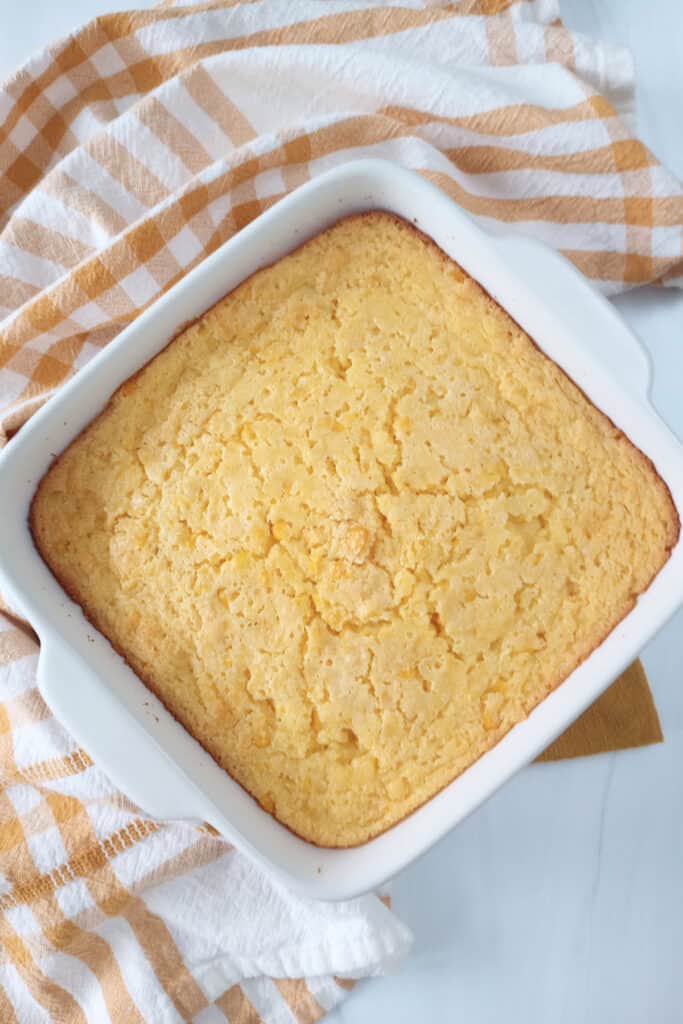 Corn bread pudding in a white baking dish next to a mustard yellow napkin and white and orange napkin. 