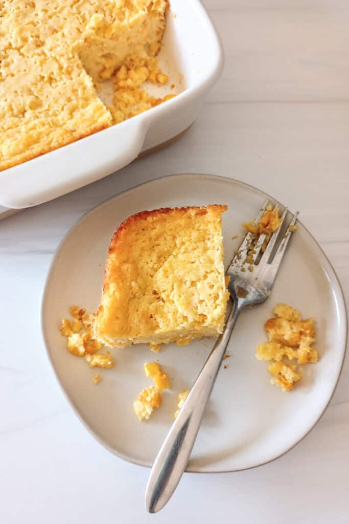 Baked corn casserole in a white baking dish next to a plate of an individual serving next to a fork. 