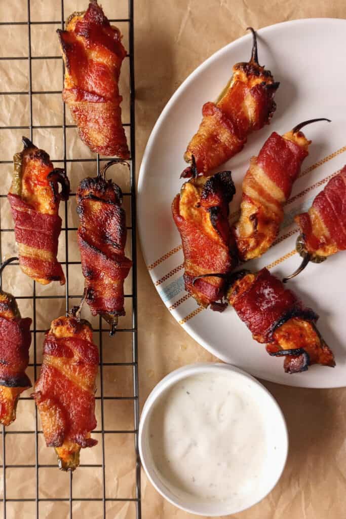 Poppers wrapped in bacon arranged on a wire rack lined with brown parchment paper. More jalapeno poppers are also arranged on a white plate. 