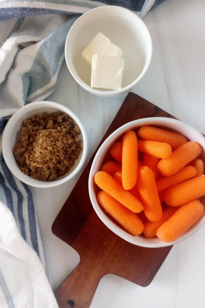 Baby carrots in a white bowl next to brown sugar and cubed butter laid out by a white table cloth. 