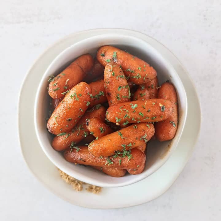 Brown sugar glazed carrots in a white bowl on a white plate next to brown sugar.