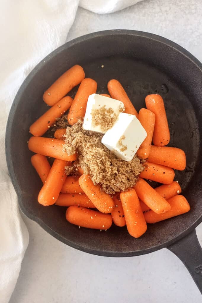Baby carrots in a black skillet topped with cubed butter and brown sugar. 
