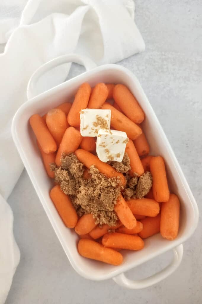 Baby carrots in a white baking dish topped with brown sugar and cubed butter ready to be baked. 