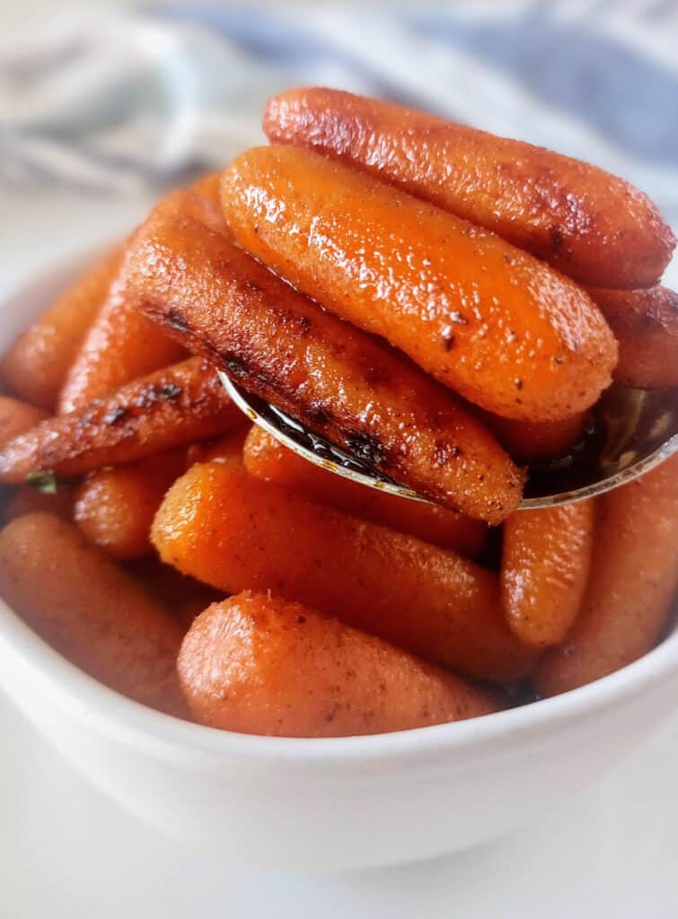 Brown sugar glazed carrots in a spoon over a white bowl. Carrots are on a white table with a white and blue cloth.