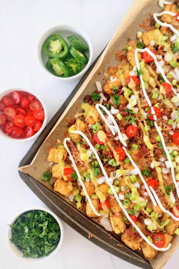 Tater tot nachos on a baking sheet next to cilantro, grape tomatoes, and green onions. 
