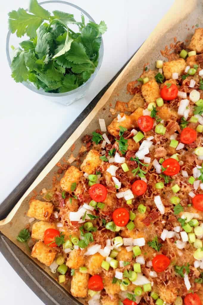 Loaded tots on a baking sheet next to a cup of cilantro leaves. 