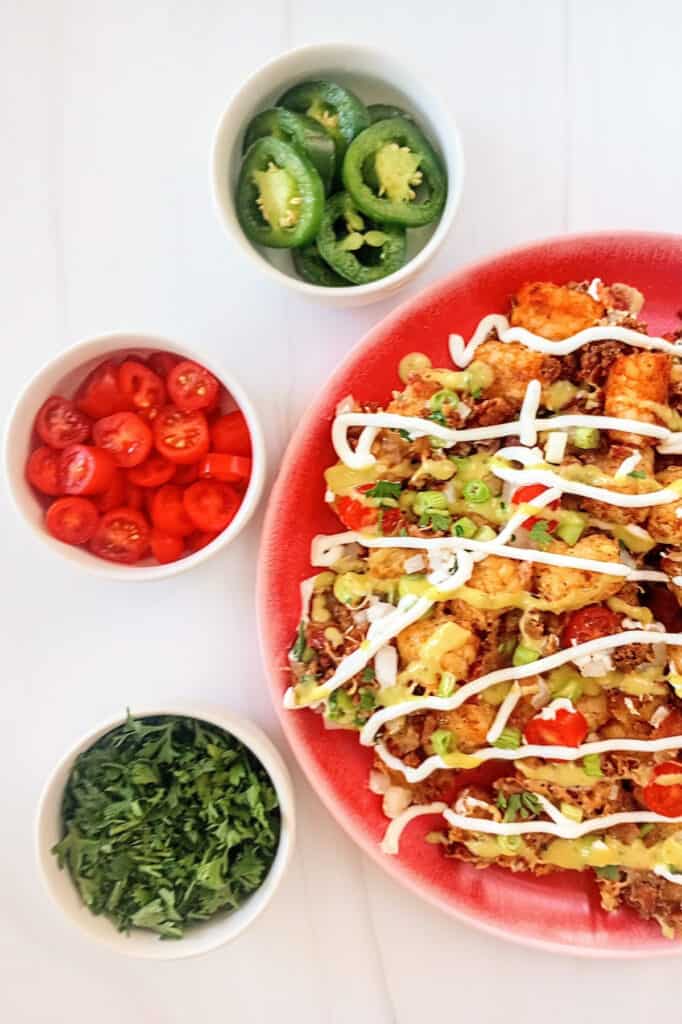 Totchos on a red plate next to bowls of cilantro, green onion , and grape tomatoes. 