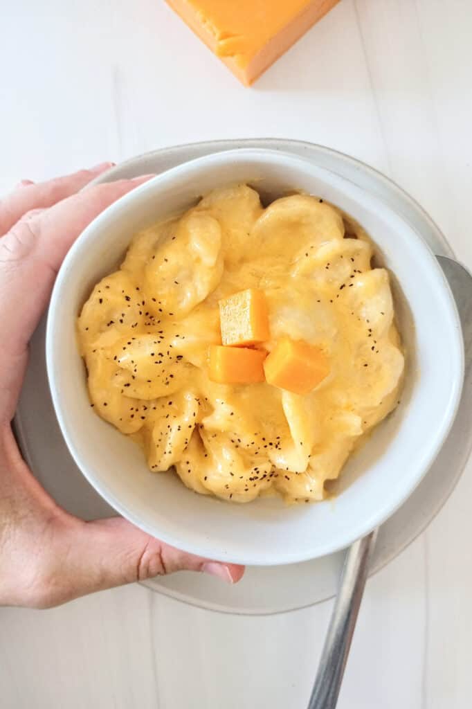 Crock-pot macaroni and cheese in a white bowl next to a spoon on a white plate. 