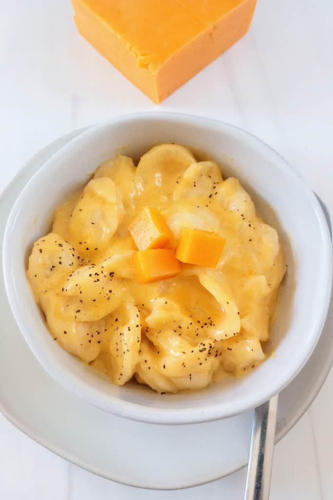 Mac and cheese in a bowl next to block cheese, topped with cubed cheese, sprinkled with black pepper. 
