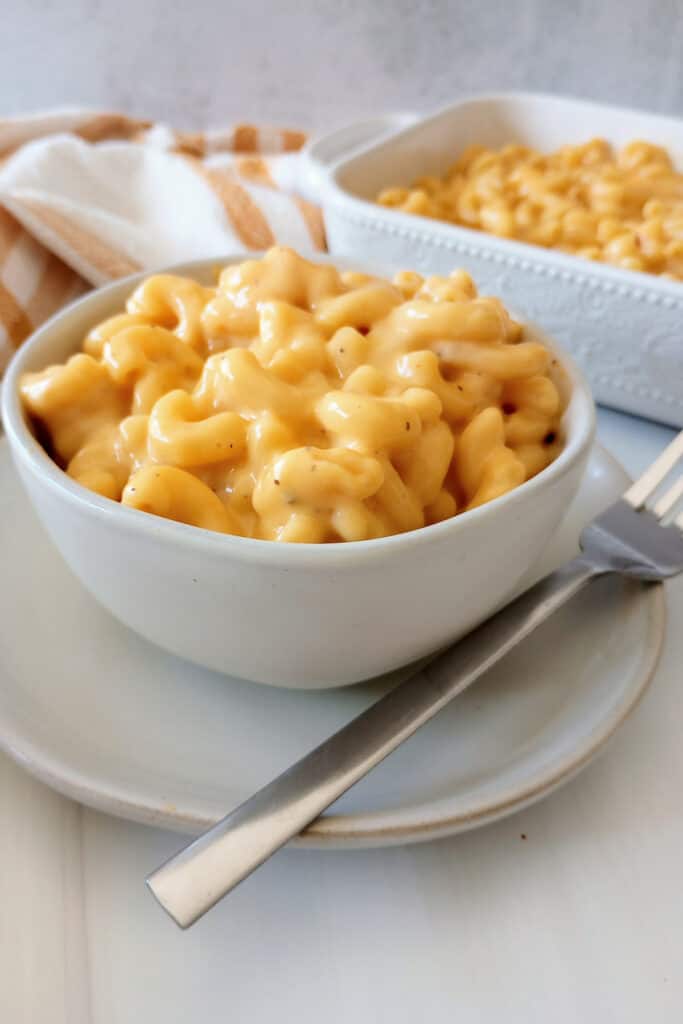 Mac and cheese in a white bowl on a white plate next to a fork. With  a casserole dish in the background.