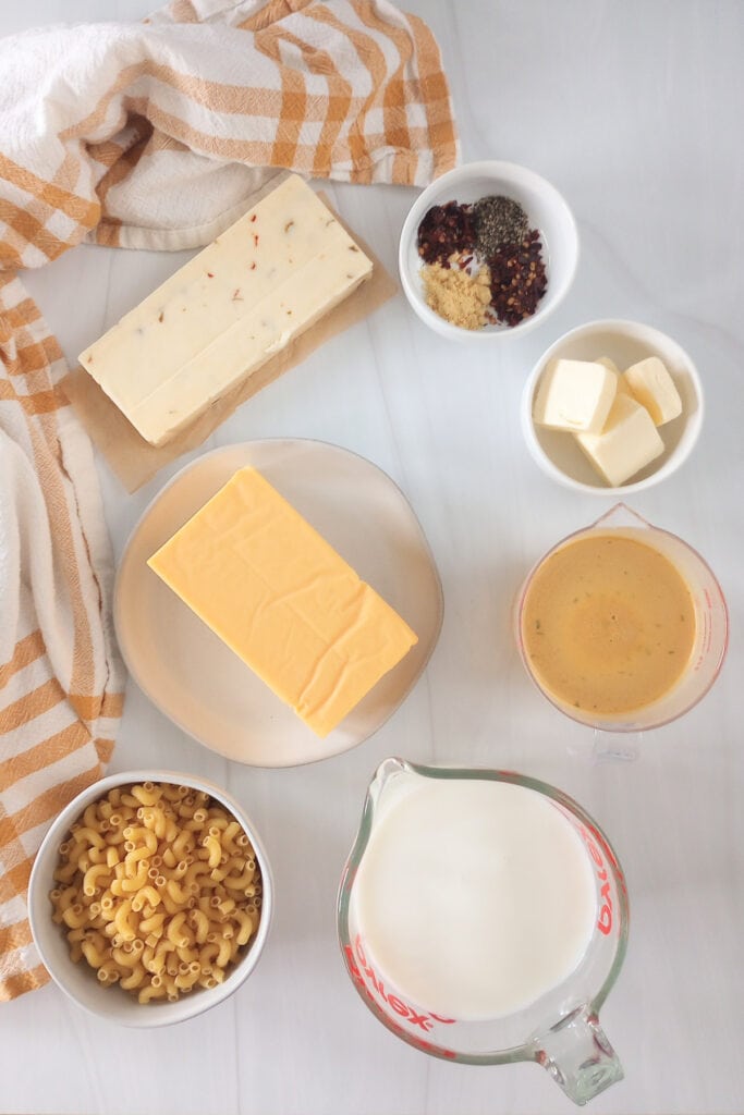 Block pepper jack cheese, milk, chicken broth, Velveeta cheese block, elbow noodles, and ground mustard, red pepper flakes, and black pepper in a white bowl. 