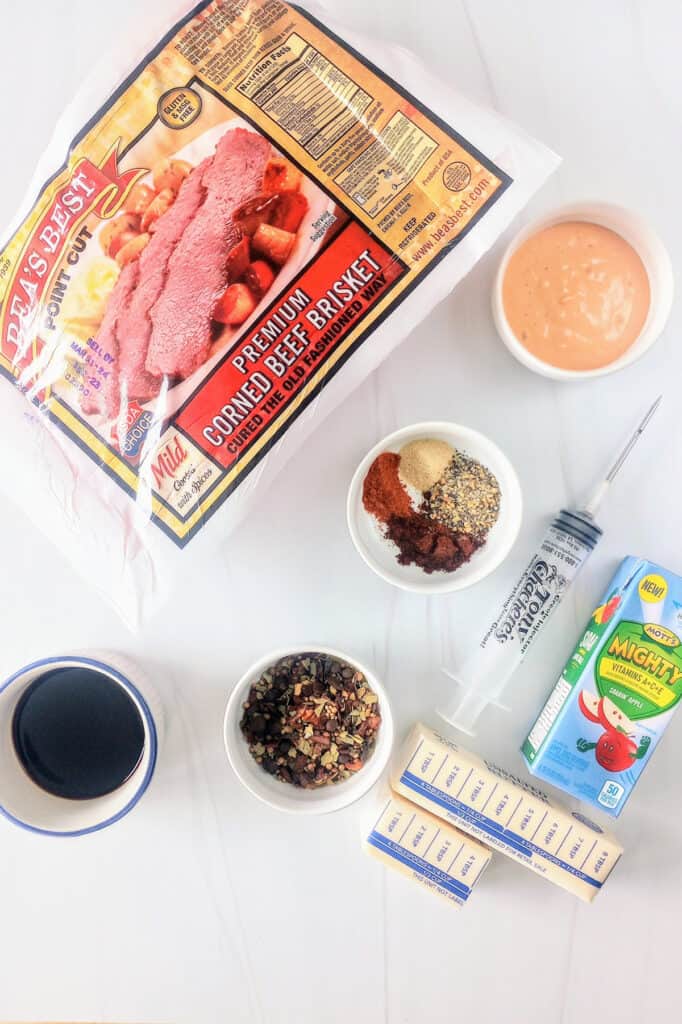 Point cut corned beef, Butter, balsamic vinegar, seasonings, meat injector, thousand island dressing, corn beef seasoning packet, and mini apple juice  laid out on a table. 