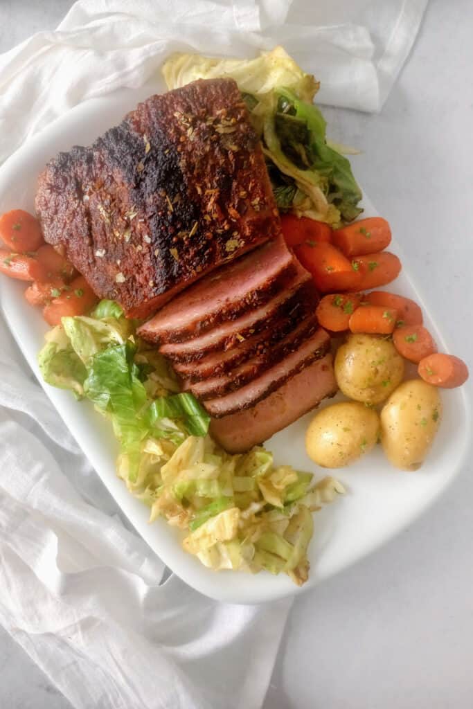 Corned beef fresh out of the oven on a white serving plate next to boiled potatoes, sauteed cabbage, and sliced carrots.