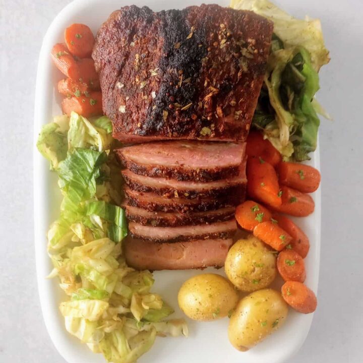 Oven baked corned beef on a white serving plate next to sauteed cabbage, sliced carrots, and boiled potatoes.