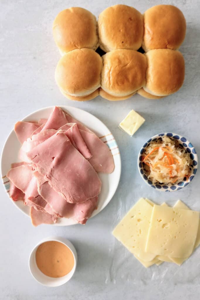 All the ingredients it takes to make Reuben sandwiches. Hawaiian rolls, thinly, sliced, deli style corned beef, Swiss cheese, thousand island dressing, and sauerkraut with thinly sliced carrots. All arranged on a grey table in white bowls or plastic wrap. 