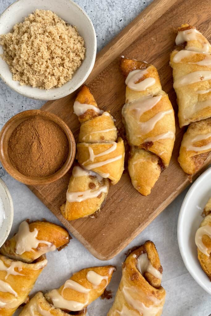 Glazed cinnamon sugar crescent rolls ready to eat, sitting on a platter next to small bowls with brown sugar and ground cinnamon.