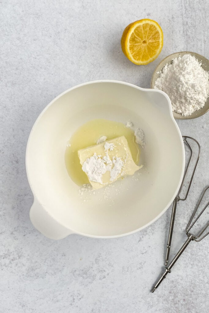 Mixing bowl with half block of cream cheese, powdered sugar and lemon juice, next to two beaters, and half a lemon.