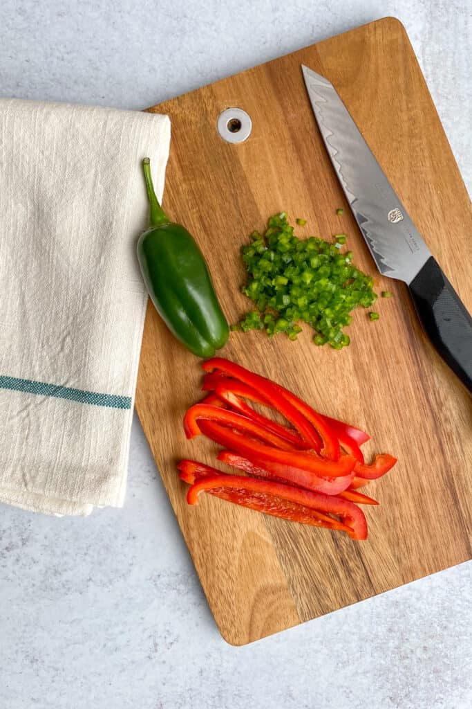 Red bell pepper and jalapeno being diced on a wood cutting board.