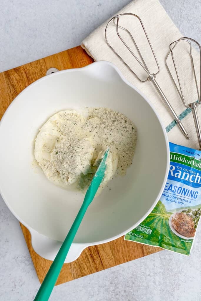 A mixing bowl that has cream cheese and ranch seasoning in it, next to a packet of ranch seasoning mix and beaters for an electric mixer.