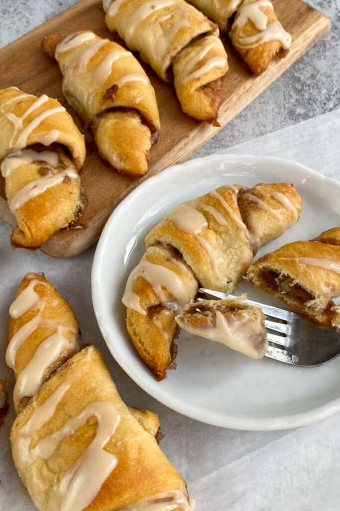 Dessert crescent rolls filled with cinnamon sugar and topped with a sweet vanilla glaze on a platter and one on a plate with a fork lifting up a gooey bite.