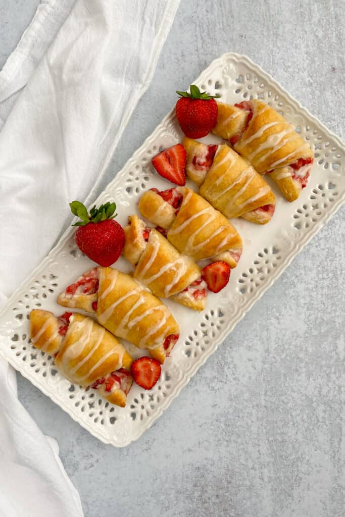 White platter with eight strawberry cream cheese crescent rolls lined up with whole and half strawberries scattered around.