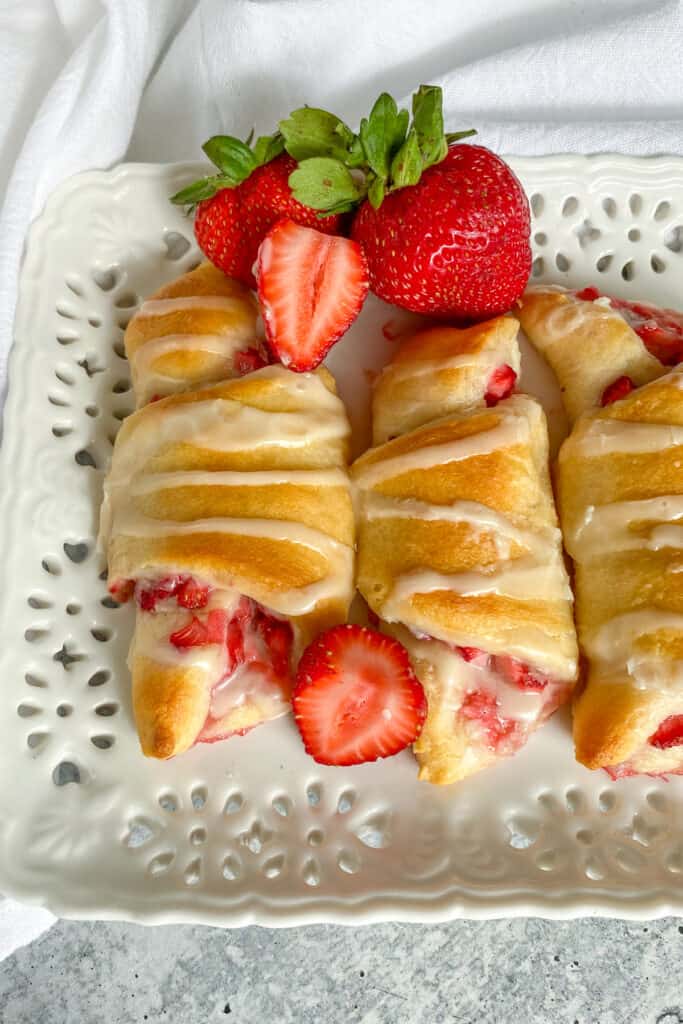 A close up view showing three strawberry cream cheese crescent rolls on a white platter with warm fillings and glaze dripping on them.