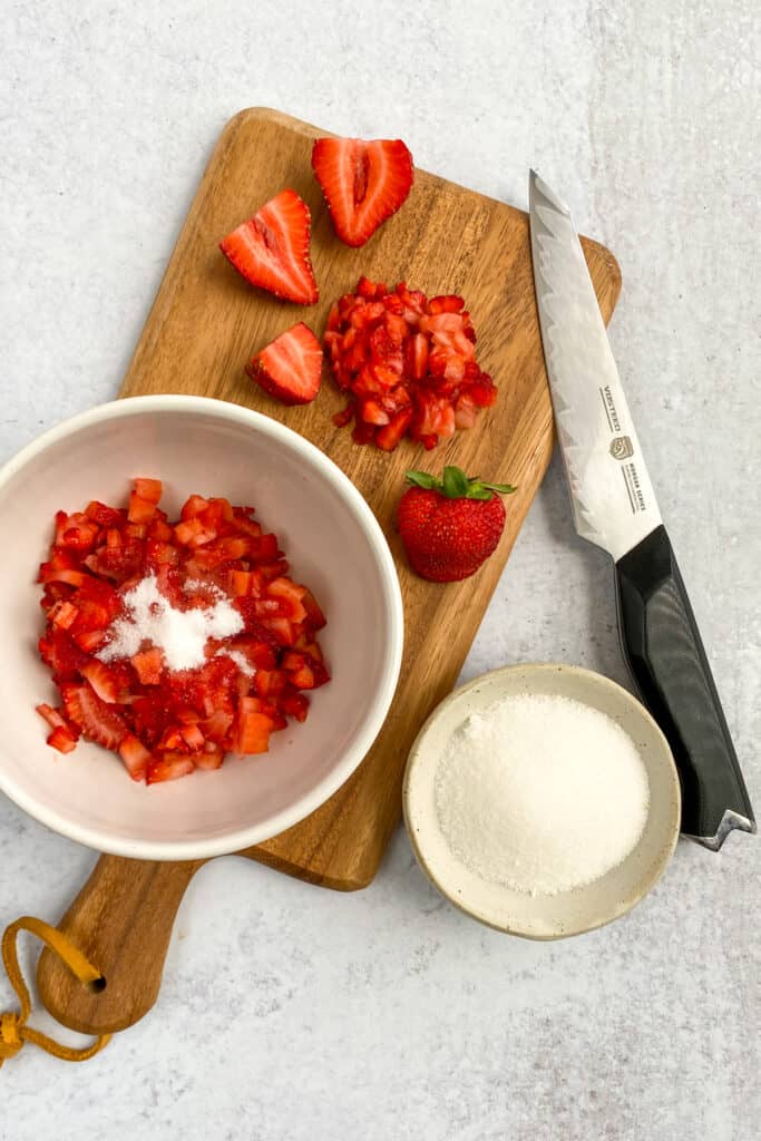 Bowl of finely chopped strawberries with sugar on top, sitting on a wood cutting board with chopped strawberries, a knife and small bowl of sugar.